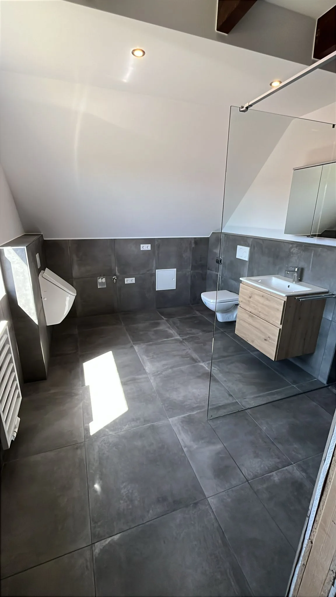 Selbes Badezimmer unter Dachschräge mit Urinal, Wand-WC, Waschtisch und Handtuchheizkörper.